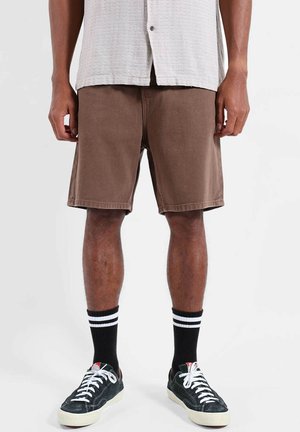 Homme portant un short marron jusqu'aux genoux, des chaussettes noires avec des rayures blanches, et des baskets noires avec des lacets blancs, debout devant un fond uni.