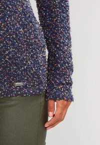 Maglione a maglia di colore blu navy con texture e fili colorati, dotato di maniche lunghe e un piccolo logo in metallo tagliato all'orlo. Abbinato a pantaloni verde oliva.