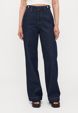 Femme portant un jean bleu foncé taille haute à jambes larges, associé à des sandales noires à bout ouvert, debout devant un fond clair uni.