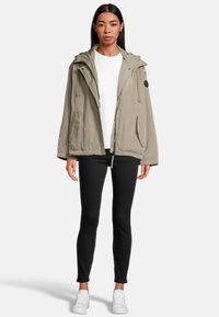 Beige Kapuzenjacke mit einem Reißverschluss vorne, zwei Seitentaschen und einer strukturierten Oberfläche. Über einem weißen Shirt und schwarzen Leggings getragen, kombiniert mit weißen Turnschuhen.