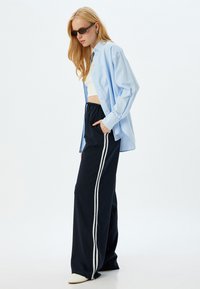 Lichtblauw overhemd met knopen, witte crop top, losse zwarte broeken met witte strepen en witte instap schoenen.