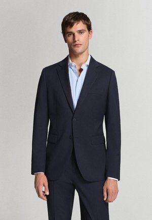 Man in een donkerblauw pak en lichtblauw overhemd staande tegen een effen lichtgrijze achtergrond.