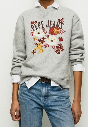 Persoon die een lichtgrijze trui draagt met de tekst "PEPE JEANS" en rode, gele en witte bloemembroidery, gecombineerd met een blauwe spijkerbroek en een wit overhemd.
