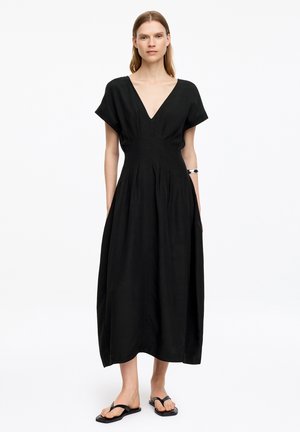 Kobieta ubrana w czarną sukienkę midi z dekoltem w serek, krótkimi rękawami i czarne płaskie sandały, stojąca na jednolitym białym tle.