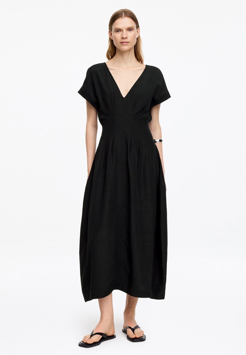 Femme portant une robe midi noire à col en V avec des manches courtes et des sandales plates noires, debout devant un fond blanc uni.