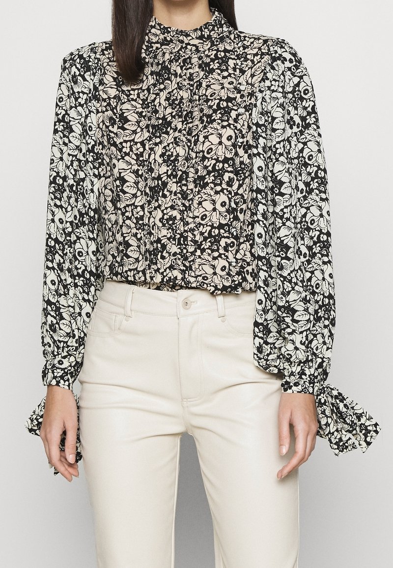 Blouse à motif floral noir et blanc avec un col haut, des manches bouffantes et des poignets froncés, associée à un pantalon beige clair taille haute.