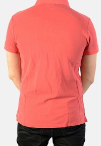Polo de manga corta en color coral con una textura suave, cuello y aberturas laterales, combinado con jeans negros. La vista trasera muestra el ajuste y la longitud de la camiseta.