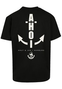 Zwarte katoenen t-shirt met "AHOI" in vette witte letters, omgeven door pijlen, met "Knut & Jan | Hamburg" en een nautische afbeelding eronder.
