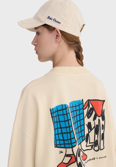 Femme aux cheveux tressés portant une casquette beige et un pull avec un graphisme coloré représentant des jambes en pantalon bleu, des chaussures rouges et un chien.