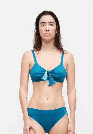 Femme aux cheveux mouillés portant un bikini bleu sarcelle avec un nœud noué sur le haut, debout devant un fond blanc uni.