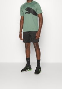 Zielona sportowa koszulka z logo czarnej pantery, w połączeniu z czarnymi szortami i czarnymi butami z zielonymi akcentami i logo.