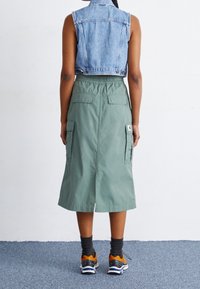 Krótka kamizelka dżinsowa zestawiona z zieloną spódnicą o średniej długości z kieszeniami cargo. Spódnica ma elastyczną talię i rozcięcie z tyłu.