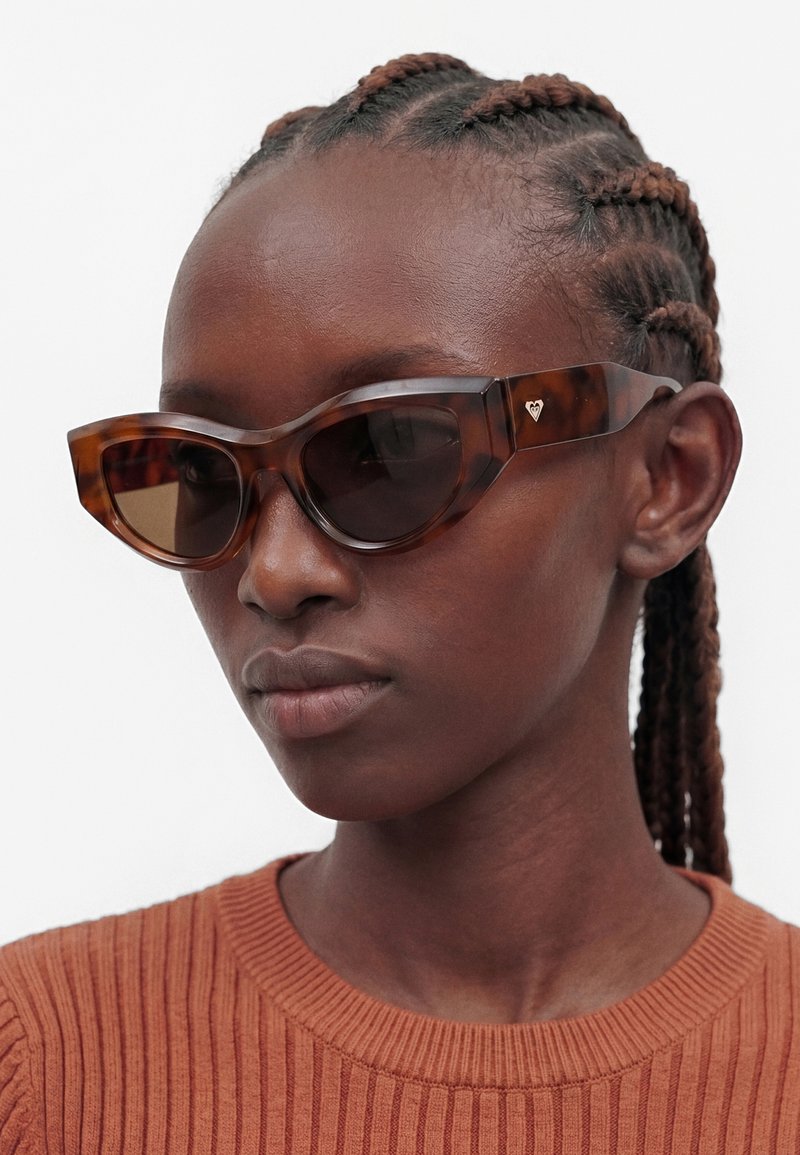 Femme aux cheveux tressés portant des lunettes de soleil œil de chat à monture écaille de tortue marron et un pull côtelé couleur rouille.