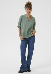 Blusa verde con texture, scollatura a V e maniche corte gatherate, abbinata a jeans a campana blu e stivaletti neri alla caviglia.
