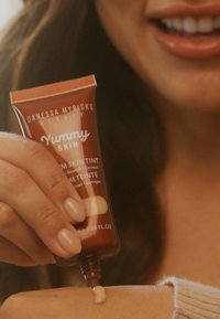 En brun tube med "Yummy Skin" hudtoner som klemmer ut en lys beige krem på en underarm med en strikket gensererm.