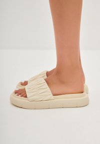 Sandales à glissière beiges avec un dessus matelassé et des semelles épaisses texturées. Style ouvert à l'avant, mettant en valeur des ongles de pieds peints en blanc.