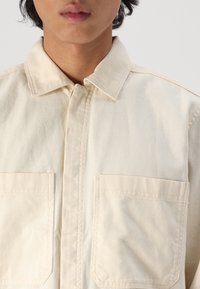 Beige, collared shirt made of woven fabric, featuring a front button closure and two patch pockets on the chest. Smooth texture.