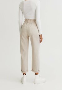 Pantalon en denim beige avec une taille haute froncée et des revers roulés. Les poches arrière sont visibles. Associé à des baskets montantes blanches.