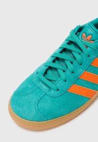 Turkio spalvos zomšiniai sportbačiai su oranžinėmis juostelėmis, tekstūruotais raišteliais ir guminiais padais. Šone yra trys išskirtinės juostelės.