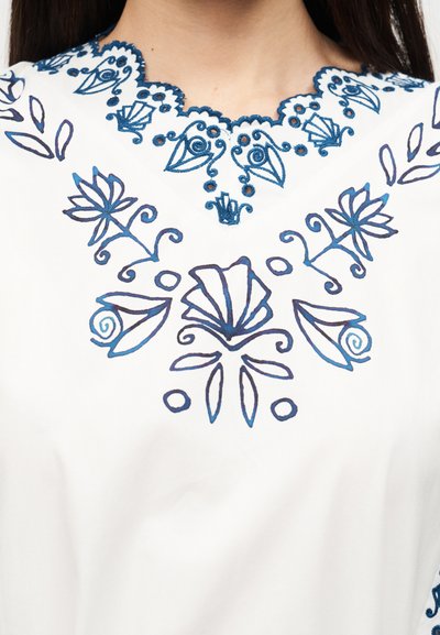 Λευκό ύφασμα με μπλε λουλουδάτη και αφηρημένη κεντήματα γύρω από λαιμόκοψη με κυματιστό περίγραμμα, που φοριέται από άτομο με σκούρα ίσια μαλλιά.