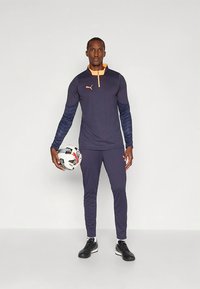 Športový set PUMA s tmavomodrým tričkom s oranžovým zipsovým golierom a dlhými rukávmi, doplnený tmavomodrými nohavicami. Model drží futbalovú loptu.