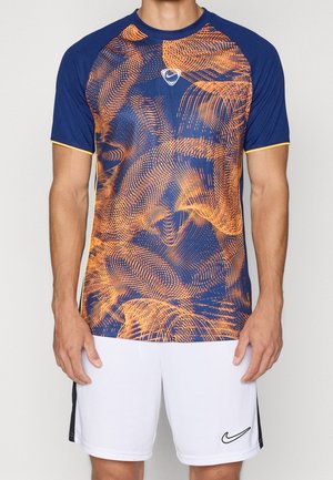 Homme portant un t-shirt de sport bleu marine et orange à motif abstrait et un short blanc Nike avec logo swoosh noir.