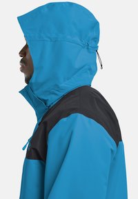 Veste imperméable bleue avec une section supérieure noire. Caractéristiques : capuche, détails zippés et texture lisse et brillante. Profil latéral montré.