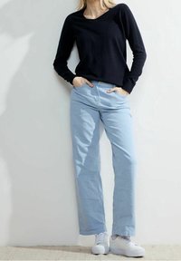 Svart långärmad topp som matchas med ljusblå raka jeans och vita sneakers, mot en neutral bakgrund.