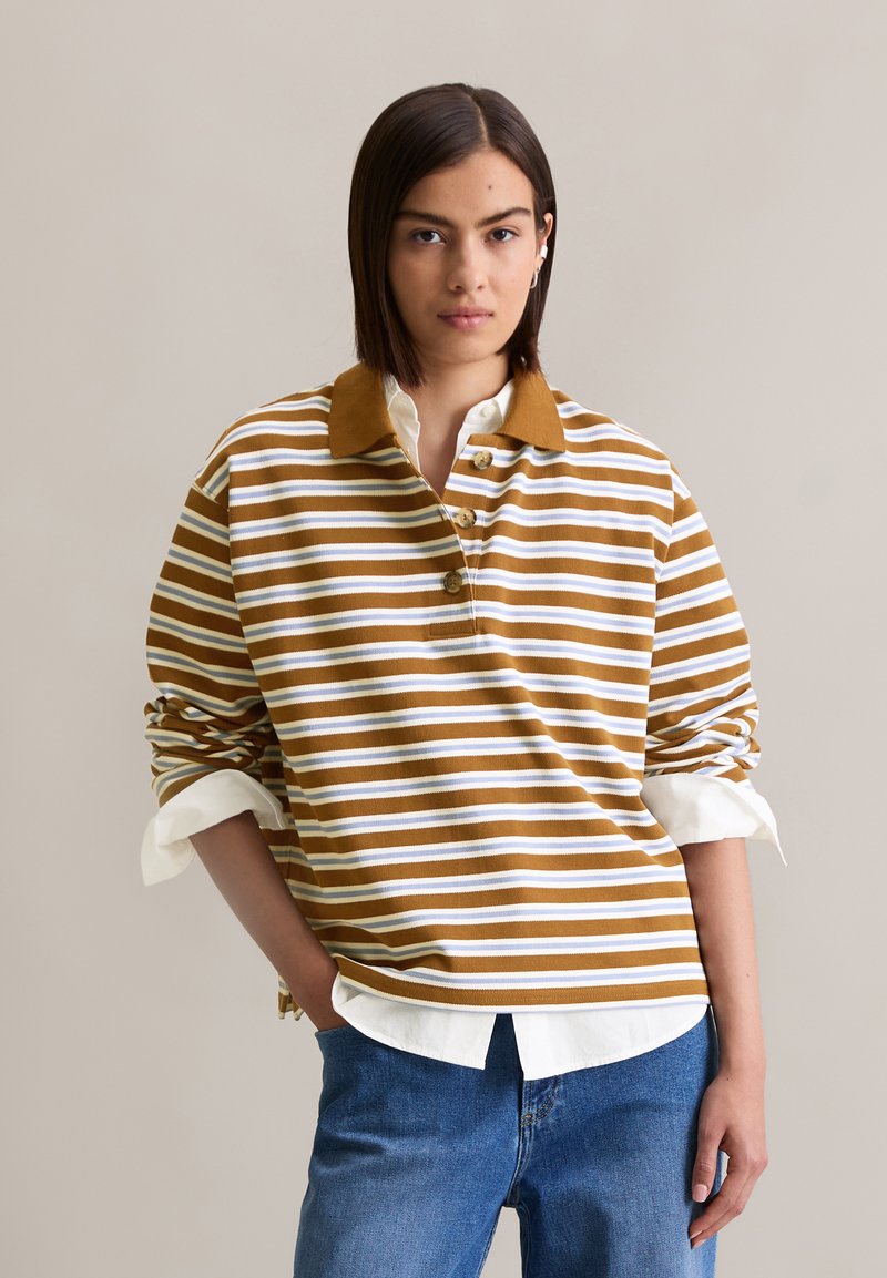 Bruine en witte gestreepte blouse met lange mouwen, een kraag en een knoopsluiting, gedragen over een witte shirt, gecombineerd met blauwe jeans.
