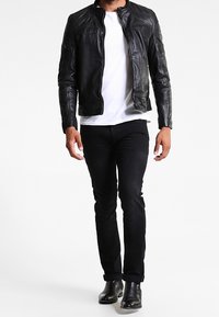 Schwarze Lederjacke mit tailliertem Design, Reißverschluss und Schulterdetails, kombiniert mit einem weißen Hemd und schwarzen Jeans.