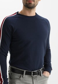 Camisa de manga larga navy con mangas raglán, con rayas rojas y blancas. Combinada con vaqueros grises y un cinturón negro. Textura suave.