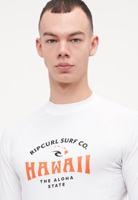 Biała koszulka rash guard z czarnym i pomarańczowym napisem. Wzór zawiera "Ripcurl Surf Co." oraz "Hawaii" z logo surfingowym, gładka tekstura, sportowy krój.
