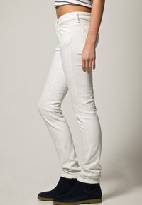 Jean slim-fit blancs avec une taille mi-haute, présentant une fermeture à bouton et à zip à l'avant, ainsi que des poches sur les côtés et à l'arrière. Associé à des bottines noires.