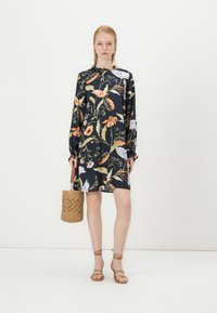 Vestido estampado com flores em azul marinho escuro, com mangas longas, corte descontraído e detalhes em botão. Modelo segura uma mala de palha trançada e usa sandálias de tiras em tom de areia.