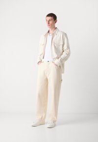 Chemise à rayures de couleur claire, portée sur un t-shirt blanc, associée à un pantalon beige ample et des baskets blanches. Détails de design minimalistes.