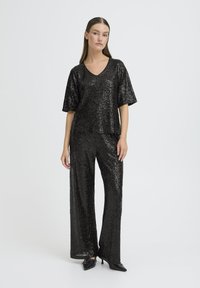 Musta paljettitoppi ja leveälahkeiset housut. V-kaulus, lyhyet hihat ja kiiltävä pinta. Yhdistetty viistoihin mustiin kenkiin.