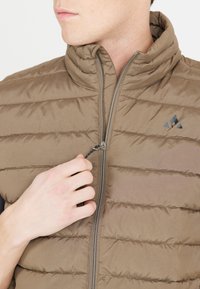 Bruine donzen gilet met diagonale stiknaden, hoge kraag en een voorkant met ritssluiting. Voorzien van een logo op de linkerborst en een trekband detail op de rits.