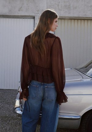 Femme aux longs cheveux porte un chemisier marron transparent et un jean bleu ample, debout à côté d'une voiture vintage argentée devant des portes de garage fermées.