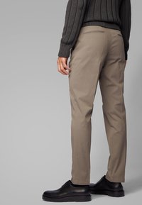Pantalon beige en coton à coupe ajustée, avec deux poches arrière et une texture lisse, associé à des chaussures en cuir noir.