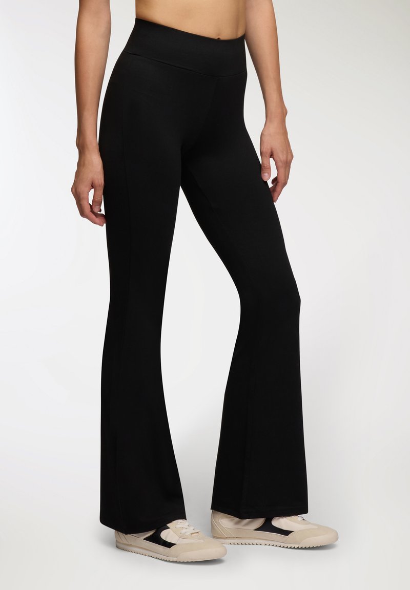 Czarne leginsy dzwonowe wykonane z miękkiego, elastycznego materiału z wysokim stanem i gładką fakturą, w połączeniu z jasnymi trampkami.