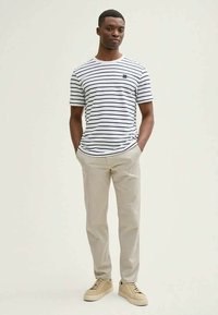 Ριγέ κοντομάνικη μπλούζα σε λευκό και ναυτικό μπλε, συνδυασμένη με μπεζ παντελόνι και μπεζ αθλητικά παπούτσια. Η μπλούζα έχει ένα μικρό λογότυπο στο στήθος.