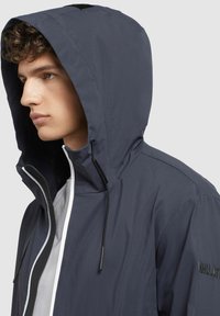 Veste imperméable navy avec une grande capuche, fermeture éclair et cordons de serrage. Elle présente une doublure intérieure blanche et un détail logo sur le côté. Tissu durable.