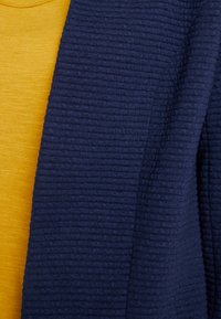 Laivastonsininen teksturoitu neuletakki, joka on päällekkäin vaaleankeltaisen paidan kanssa. Neuletakissa on ribbitekstuuri, joka tuo ulottuvuutta ja syvyyttä.