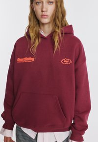 Bordó kapucnis pulóver első zsebbel, narancssárga "Túlzott gondolkodás" felirattal és egy kis felhő grafikával. Puha anyag és kényelmes szabás.