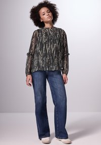 Schwarze durchsichtige Bluse mit weißem gepunktetem Muster, lange Ärmel mit Rüschensäumen, kombiniert mit blauen Schlagjeans und hellen Schuhen.