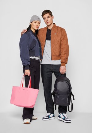 Bolso tote rosa con asas rojas, chaqueta bomber cortada en azul marino, chaqueta acolchada marrón, jeans oscuros y mochila negra con acentos blancos.