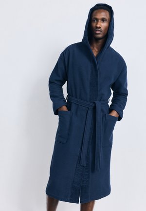 Uomo che indossa un lungo accappatoio con cappuccio blu navy con tasche e cintura, in piedi con le mani in tasca davanti a uno sfondo semplice.