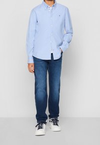 Camisa de botones azul claro con mangas largas, jeans de mezclilla y zapatillas blancas. Presenta un pequeño logo en la camisa y un corte relajado.