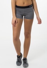 Sportowiec ubrany w szare szorty Nike i czarno-szare buty do biegania Nike, stoi z nogami lekko rozstawionymi na tle białym.