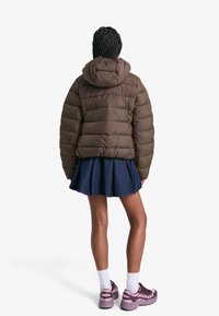 Rjava napihnjena jakna s kapuco, prešita tekstura, v kombinaciji s temnimi kavbojkami in rožnatimi supergami, ki prikazuje plastičen sproščen videz.